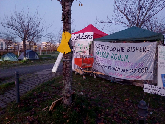 Am Rand des Bürgerparks in Köln-Kalk stehen Pavillons der Mahnwache für die "Sündi 4", daran hängen Banner: "Weiter so wie bisher: Wälder roden, Aktivistis verurteilen, leere Versprechen auf der COP! fuck RWE! Sündi lebt!"; im Hintergrund zwei Zelte auf einer Wiese mit kahlenLaubbäumen, weiter hinten eine Häuserfront
