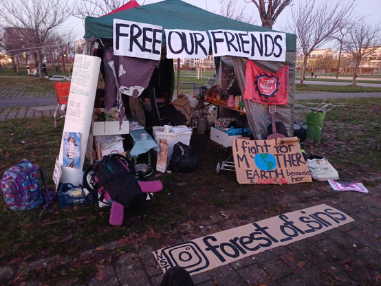 Mahnwachen-Pavillon mit Fahnen und Bannern: "FREE OUR FRIENDS", "Fight for Mother Earth" und "(Insta) forest_of_sins"; im Hintergrund kahle Laubbäume im Park
