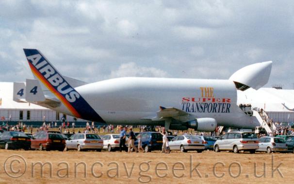 Side view of a very large, twin engined jet freighter with an over-sized cargo hold on top of an already pretty big body, with the front of the fuselage hinged upwards, giving access to the cargo area.
The plane is mostly white, with very large titles "The Super Transporter" on the side of the upper fuselage, each word in yellow, red, and blue, respectively.
The black registration "F-GTSD" is on the lower rear fuselage.
The rear fuselage and tail have a thick, diagonal blue band with smaller red, orange, and yellow bands towards the rear, with the white word "Airbus" overlaid on the blue stripe.
That same logo, minus the text, is repeated on the wingtip fences on the ends of the wings.
There are large end plates on the horizontal stabiliser, with the number "4" on both the inner and outer faces.
There is a large, white fillet extending the tail to meet the massively extended upper fuselage, that fuselage extension eating up a large chunk of the normal tail.
There is a white set of mobile airstairs parked leading up to the open freight deck, with people climbing the stairs, and others admiring the view from the open deck.
Cars are parked on grass in the foreground, with people wandering around, and more people in the background, in front of a large white building which stretches across the whole frame.
Fluffy grey clouds with hints of blue peeking through fills the rest of the frame.