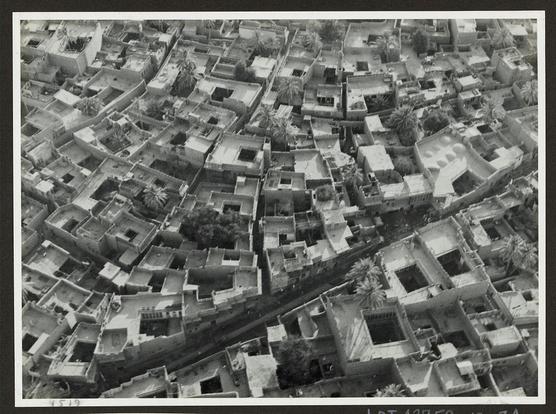 The image shows an aerial view of a densely packed urban area with numerous closely built buildings. The architecture appears traditional, featuring flat roofs and courtyards within the structures. There are several trees scattered throughout the scene, likely providing shade in this arid environment. A prominent street runs through the center of the photograph, dividing the neighborhood into two distinct halves. The image is monochromatic, suggesting it might be an older photograph or taken with black-and-white film. It's labeled as "Baghdad," indicating that the location depicted is within Baghdad city in Iraq.