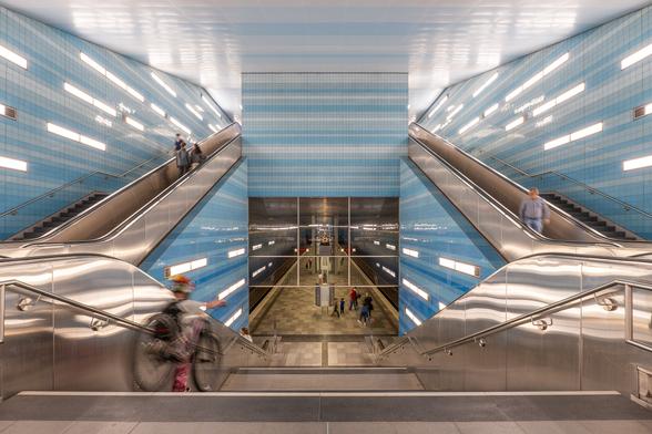 Blick von der in Blautönen gestalteten Treppenanlage auf den Bahnsteig. Ein Fahrgast trägt sein Fahrrad die Treppe herunter. Auf dem Mittelbahnsteig befinden sich wartende Fahrgäste.