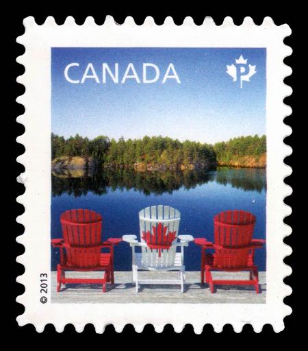 A postage stamp features 2 red and one white (with a red maple leaf)Adirondack-style chairs arranged to look like a Canadian flag, on a wooden dock overlooking a calm lake surrounded by lush green trees. The words "CANADA" appear in the upper left corner along with a Canadian leaf, and a copyright symbol is visible at the bottom.