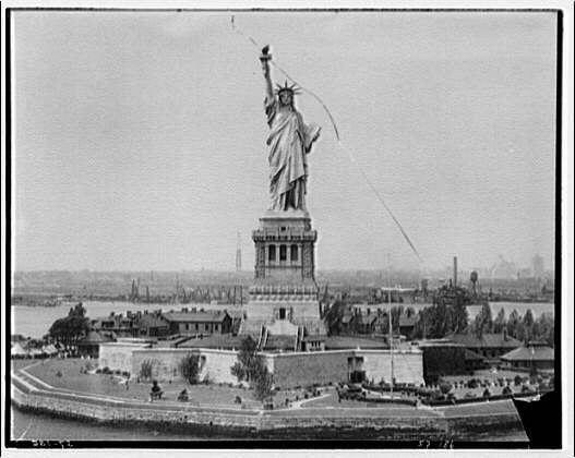 The image shows a black and white photograph of the Statue of Liberty, an iconic monument located in New York Harbor. The statue is depicted with its full stature, standing on a pedestal surrounded by water with boats visible in the background. There are people at the base of the statue, giving context to the scale of this colossal sculpture. Smokestacks can be seen across the harbor, suggesting industrial activity near the port area. This photograph appears to have been taken from an elevated perspective, providing a comprehensive view of both the Statue and its surroundings during what seems like late 19th or early 20th-century era based on the image quality and style.