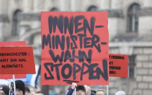 Eine Protestszene zeigt verpixelte Demonstrierende mit einem roten Schild mit schwarzem Schriftzug „Innenministerwahn stoppen!“