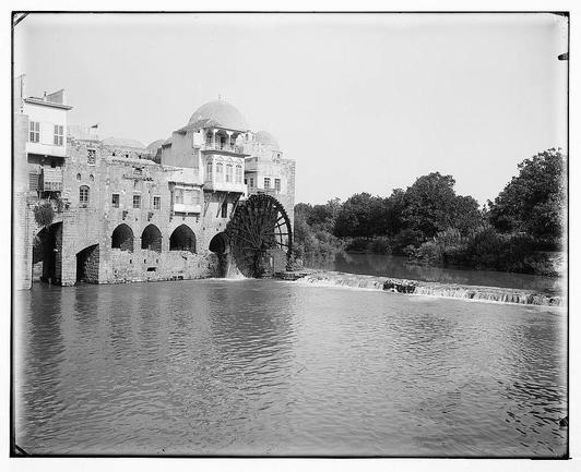 The image is a black and white photograph depicting an ancient watermill alongside a river. The mill has large, circular stone wheels that are partially submerged in the flowing water, indicating its use for generating power or processing grain. Adjacent to the milling building, which features arched doorways and windows with protective bars, there's another structure topped by a dome-like roof resembling an Islamic architectural style commonly found in historical Middle Eastern regions.

The surrounding environment includes lush greenery with trees lining both sides of the riverbank, suggesting that this location is near nature. The water appears calm despite some flow from small cascades or rapids visible on its surface. There's no indication of modern development around the site, which gives it a timeless quality as if frozen in history.

The photograph has an edge-to-edge border and seems to be mounted or framed for display purposes. A reference number "691e" is printed at the top right corner within the image frame, likely indicating its cataloging information within a collection of photographs. The photo's monochromatic tone adds to the nostalgic atmosphere and emphasizes textures such as stone masonry on the buildings and river water ripples.

The context provided indicates that this photograph could be part of an archive from Emeric and Edith Matson regarding their travels, specifically related to nature (as deno [...]