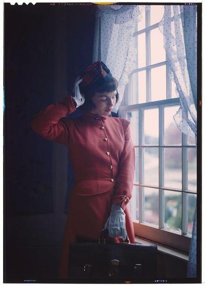 This color transparency photograph features a woman dressed in a vibrant red suit, complemented by a matching hat adorned with feathers. She holds her head and appears to be adjusting or looking at something on the side of it. The ensemble is completed with white gloves and she stands next to an open window that reveals daylight outside. Her gaze directs downwards towards what seems like a briefcase in her hand.

The setting suggests a moment captured indoors, possibly within a residential space given the presence of sheer curtains adorned with polka dots partially framing the large glass pane through which natural light filters into the room. The overall ambiance evokes an atmosphere reminiscent of mid-20th-century fashion and lifestyle photography that may have been used for magazine features or personal keepsakes.

The photograph is part of a collection by Toni Frissell, an American photographer known for her work spanning various themes including portraiture and landscapes during the early to mid 1940s. The specific date range provided suggests these were likely taken between approximately 1925-1970 but could potentially be within this time frame with some variation.

Frissell's oeuvre is characterized by a dynamic approach that bridged different photographic styles and subjects, including notable portraits of influential figures such as Jacqueline Kennedy Onassis. The collection from which this image originates disp [...]