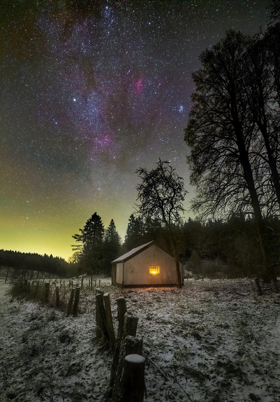 A light shines in a cottage window, bare trees behind, the Milky Way above, in a snowy setting.