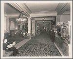 An old photograph depicting a scene from the Raleigh Hotel's lobby, featuring men seated and standing around. The room is adorned with patterned carpeting, furniture such as chairs, tables, and decorative elements like chandeliers on columns. One man can be seen sitting in the foreground reading a newspaper while others are engaged in various activities within the space.