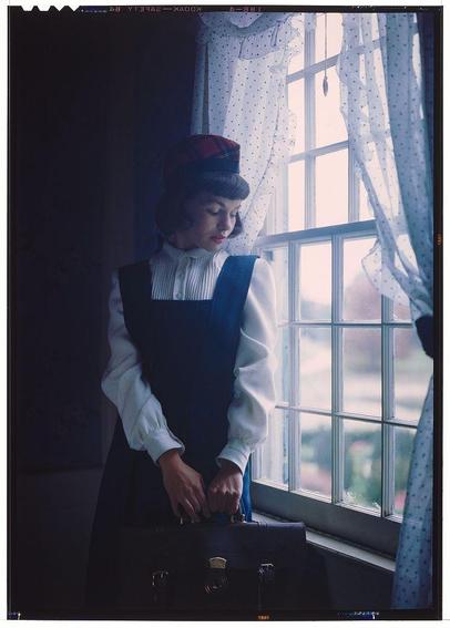 a woman standing near a window opening, wearing a white shirt with high-neck collar and dark vest over it. She is also dressed in a red hat which she appears to be adjusting or placing on her head as she looks down towards the suitcase that rests next to her. The room's lighting suggests either dawn or dusk due to light from outside shining through the window onto the sheer curtains, creating dappled patterns of light and shadow within the interior space