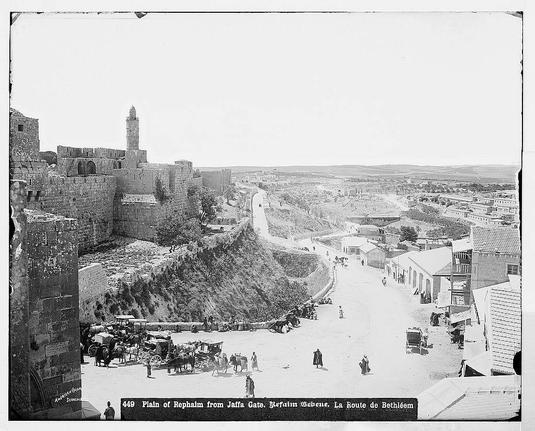 The image depicts a historical black and white photograph of the Plain of Rephaim from Jaffa Gate in Jerusalem. The view shows an expansive urban landscape with ancient stone walls, structures, and a prominent minaret-like tower indicative of Islamic architecture. A busy street scene unfolds below, bustling with people walking or standing around various small shops and market stalls lined along its length. There are horse-drawn carriages suggesting the era is likely late 19th to early 20th century. The background reveals hills and a wide expanse leading towards Bethlehem's town center. Annotations in French indicate different street names, such as "La Route de Bethléem," which translates to "The Road to Bethlehem." This historical snapshot captures daily life within an urban setting surrounded by ancient fortifications and offers insight into the architectural styles of that period.