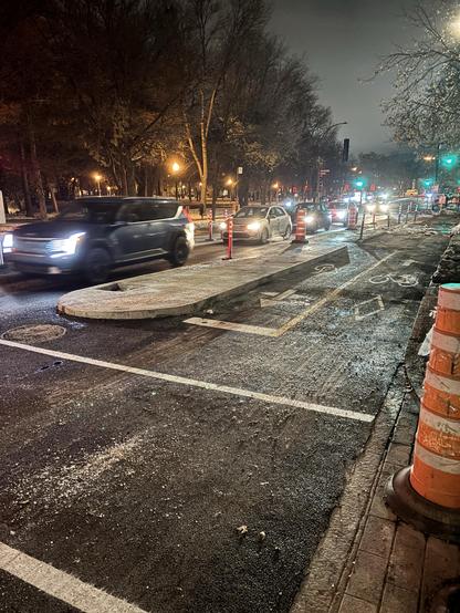 Une scène nocturne montrant des voitures circulant sur une route en travaux. Des cônes de signalisation et des barrières sont présents, et des lampadaires éclairent la zone. Des arbres bordent la rue et une piste cyclable est visible sur le côté, séparée par un îlot en béton.