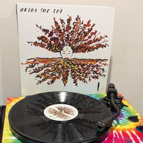 Una imagen muestra un disco de vinilo negro girando sobre un tocadiscos con un llamativo patrón tie-dye de colores rojo, naranja, amarillo, verde y azul. Detrás del tocadiscos, se alza la portada del álbum. La portada es blanca y presenta una ilustración central de un rostro estilizado y pálido con los ojos cerrados y marcas doradas en la frente. De este rostro emanan grandes masas de formas onduladas y fluidas en tonos marrones, naranjas y dorados, que se extienden tanto hacia arriba como hacia abajo, asemejándose a cabello exuberante o corrientes de energía. En la esquina superior izquierda de la portada, se lee el título del álbum: "DRINK THE SEA".