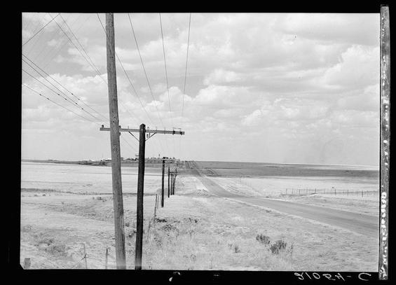 The black-and-white photograph displays a desolate landscape with an open field stretching towards the horizon. A weathered telephone pole stands prominently in the foreground, supported by multiple wooden posts and wires extending into the distance. These lines lead to additional poles further down the road, creating a pattern that draws attention along the length of what appears to be a rural highway. Sparse vegetation dots the landscape on either side of the unpaved roadway, which curves slightly as it moves away from view.

In the background, there is an expansive horizon where faint outlines suggest structures or buildings at some distance but they are not distinctly visible due to their small size and the hazy atmosphere. The sky above is filled with clouds, indicating overcast weather conditions that contribute to a somber mood in this rural scene.

The watermark "810R4 - C" suggests it may be cataloged within a larger collection or series of photographs by Dorothea Lange for her work related to the Great Plains and highway north of Amarillo, Texas.
