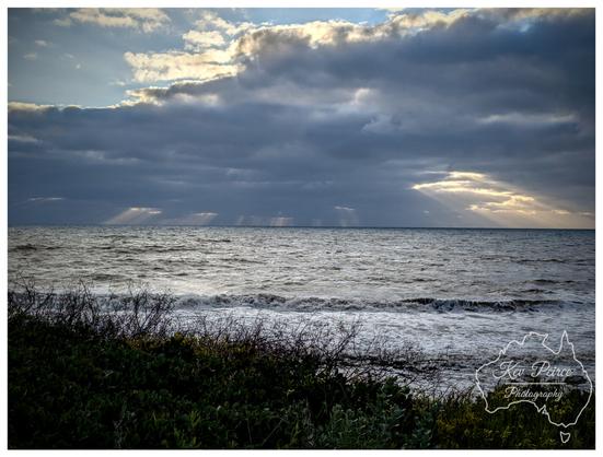 A dramatic seascape photograph taken on the Bunbury coast. The foreground features dark green, dense coastal scrub and bushes. The mid ground is a choppy, dark ocean with white breaking waves rolling towards the shore.  The horizon is visible under a vast, moody sky dominated by heavy grey clouds, with bright pockets of sun breaking through on the left and right, casting distinct light rays onto the water.