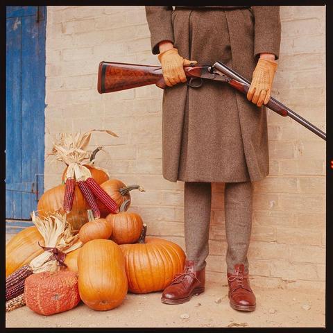 The image depicts a person standing in front of a pile of seasonal vegetables and corn, holding an old-fashioned rifle. The individual is dressed in a brown coat, gloves, pants, shoes, which indicate traditional or possibly rural attire suitable for colder weather.

In the background, there's a white brick wall that might suggest an outdoor setting near a farmstead or country home. A blue door can be seen to the left side of the frame adding contrast and depth to the composition. The person is holding the rifle with both hands, positioned as if readying it for use which could imply they are engaged in hunting activities.

The seasonal vegetables include different types of pumpkins, corn on the cob, dried maize ears among others suggesting harvest time. This image evokes a sense of rustic life and simplicity often associated with rural America during certain seasons or holidays such as Thanksgiving. It's worth noting that this photograph is part of Toni Frissell’s collection reflecting her focus on capturing everyday American lives across various conditions including economic hardships like the Great Depression.

This particular shot, titled 'Shooting Fashions - Mrs. Robert Me Leau', was taken in Philadelphia in 1963 and reflects a period when fashion trends were influenced by wartime austerity measures which led to more practical clothing styles for both men and women. It is an example of how photography can be used as h [...]