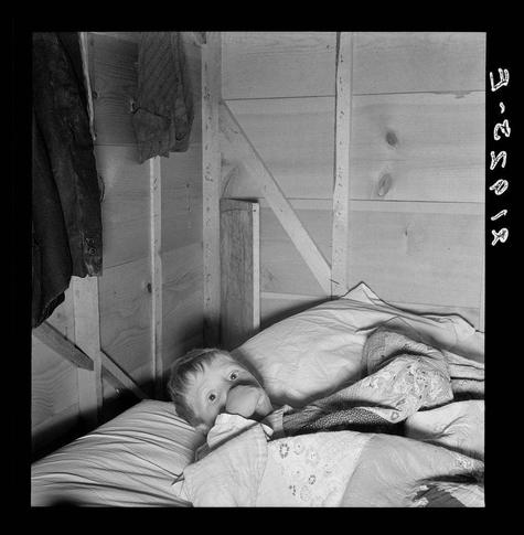 This black and white photograph captures a moment of rest or illness in what appears to be a modest, rustic living space. A child is lying on his side atop a bed covered with patterned quilts, one featuring lace details. The bedding suggests warmth but also shows signs of wear. Hanging clothes above the headboard imply that this room serves multiple functions beyond sleeping, possibly as a makeshift laundry area or general storage.

The setting seems spartan and lived-in, with wooden walls showing visible imperfections like nail holes and knots, indicating either intentional simplicity or economic constraints on construction materials. In one corner hangs what appears to be a dark coat, while the other side reveals more hanging garments, hinting at a close quarters lifestyle where space is maximized.

There's an air of quiet resilience in this snapshot: despite apparent poverty implied by the condition of their surroundings and belongings, there’s also a sense of continuity with family life—implied through objects like clothing. The scene may evoke feelings related to themes such as hardship, displacement, or rural living during challenging times for families.

Details hint at specific conditions—the photograph is credited to Dorothea Lange, known for her work documenting the Great Depression and its impact on American society; it was likely taken in Oregon by a mobile unit of FSA. Lange's images often focused on the dig [...]