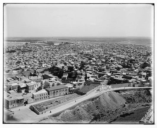 This black and white aerial photograph depicts a densely packed urban area with numerous buildings closely situated to one another. The architecture suggests an older, possibly historical cityscape, characterized by rectangular blocks of buildings with flat roofs. There are notable structures that appear larger or differently designed than the surrounding constructions, potentially indicating public or religious significance such as mosques or churches.

In the foreground, a street winds through what seems like lower residential zones, and open spaces can be seen alongside it, possibly gardens or small communal plots. To one side of this area is an elevated ground leading up to higher-lying buildings with domes that are indicative of Islamic architecture. The city spreads out towards the horizon under a hazy sky, suggesting either early morning mist or arid atmospheric conditions typical for regions like those in the Middle East.

The photograph carries signs of age and wear, including creases on its top edge, hinting at its historical value as part of the Eric and Edith Matson Photographs collection. The image appears to be from a time period between 1898 and 1946, likely capturing life in Aleppo during that era before extensive modernization or conflicts altered its appearance.

The absence of people suggests either an elevated vantage point or timing when the streets were not bustling with activity, possibly emphasizi [...]