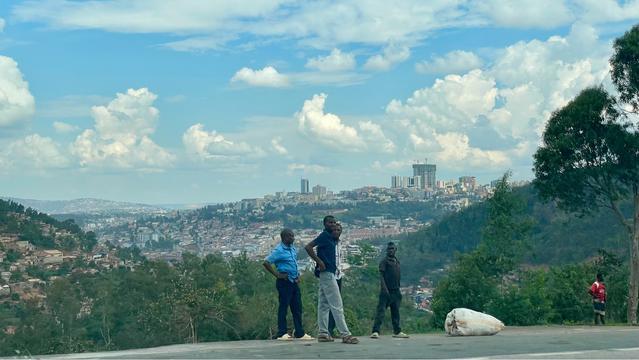 A wide view overlooking Kigali with rolling green hills and dense neighborhoods stretching across the landscape. Several men stand at an overlook in the foreground, casually gathered and looking out over the city. Tall modern buildings rise in the distance under a bright sky filled with large white clouds.