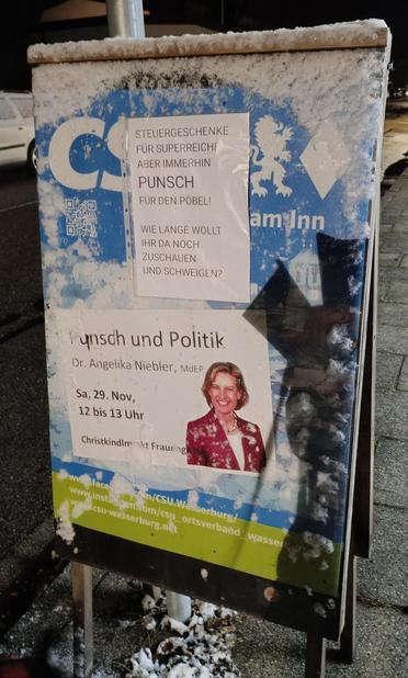 Ein Foto eines Wahlplakats, das auf einem Metallständer steht. Das Plakat hat einen dunkelblauen Hintergrund mit einem hellen, schneeflockenähnlichen Muster. Auf dem Plakat befindet sich ein Porträt einer Person mit braunen Haaren und einem dunklen Blazer. Darunter steht Text: „Punsch und Politik. Dr. Angelika Niebler, MdEP. Sa. 29. Nov. 12 bis 13 Uhr.“ Am oberen Rand des Plakats steht: „Steuergeschenke für Superreiche – aber immerhin Punsch für den Pöbel!“ und am unteren Rand „Christkindl Markt Frauengasse“. In der unteren rechten Ecke befindet sich eine Website-Adresse: „www.niebler.com/CSU-Wasserburg“ und „www.csu-wasserburg.net“.