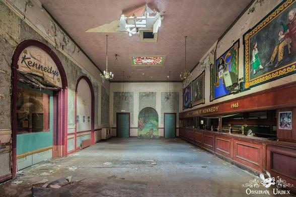 The large, derelict room has classical architectural features, peeling paint, and signs for the "Kennedys" bar and the year "1963" visible on the right wall. T