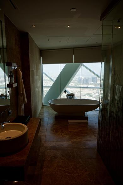 Ein luxuriöses Badezimmer mit einer modernen Badewanne, einem runden Waschbecken und einem großen Fenster mit Blick auf die Stadt. Zur Ausstattung gehören Marmorfliesen, ein an der Wand hängender Bademantel und eine Glasabtrennung für den Duschbereich.