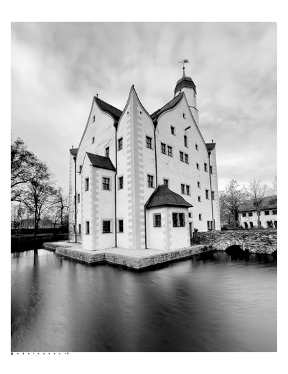 Aufnahme in schwarzweiß, Seitenansicht vom Wasserschloss Klaffenbach (Stadt Chemnitz, Deutschland), aus der Zeit der Renaissance, heller Putz, kastenförmiger Bau mit kleinen Fenstern und geschwungenen Giebelformen, plus außermittigen Turm, gefüllter Wassergraben, durch Langzeitbelichtung verschwommen, links kahle dunkle Bäume, hellgrauer Himmel darüber