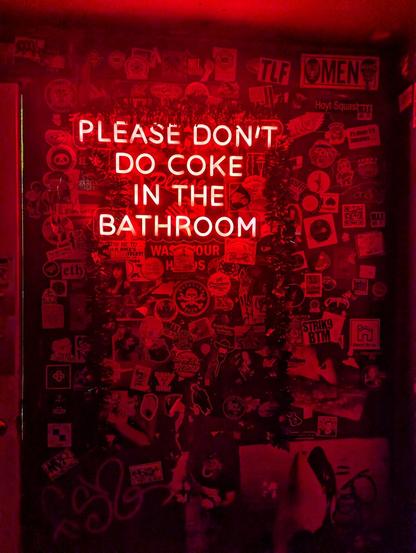 The image displays a neon sign in red that reads, "Please don't do coke in the bathroom," surrounded by an array of stickers and graffiti on a dark wall.
