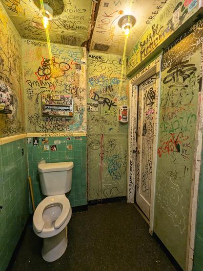 The interior of a compact, intensely graffitied restaurant bathroom cubicle. Every available surface, including the walls, ceiling, and door, is covered in a dense layer of colourful graffiti, tags, drawings, and stickers, creating a vibrant and chaotic atmosphere.

The toilet is positioned in the left corner, with green tiles covering the lower half of the walls. The upper walls and ceiling are painted and filled with handwritten messages, artistic sketches, and various markings. The lighting is provided by two overhead bulbs, casting a warm glow on the eclectic decor.

The door to the cubicle is also heavily adorned with graffiti, and a small vent is visible near the ceiling. The overall ambiance is gritty and urban, reflecting a space that encourages self-expression and artistic freedom.