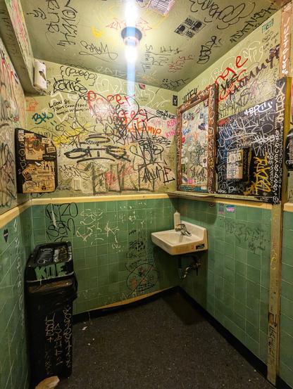 The interior of a compact, edgy restaurant bathroom, heavily adorned with graffiti and stickers. The walls and ceiling are covered in a dense layer of colourful tags, drawings, and messages, creating a vibrant and chaotic atmosphere.

The sink area features a white porcelain sink with a mirror above it, both of which are surrounded by graffiti and various stickers. To the left of the sink, there is a black bin with more graffiti on it. The lower half of the walls is tiled in green, while the upper half and ceiling are painted and filled with an array of handwritten notes and artistic markings.

The lighting is provided by a single overhead bulb, casting a bright light on the eclectic and creative decor. The overall ambiance is gritty and urban, reflecting a space that encourages self-expression and artistic freedom.