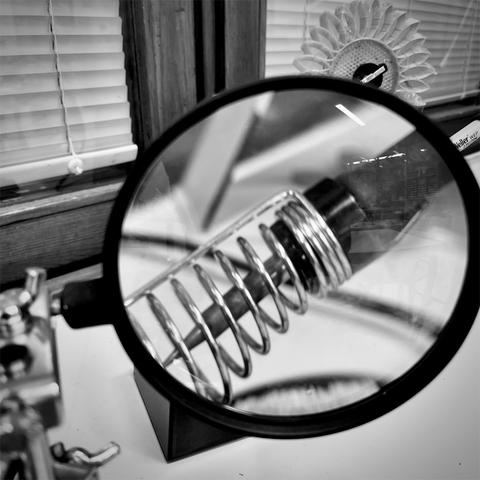 A close-up of a magnifying glass focusing on a metal spring holder of a soldering iron, with blurred background elements in black and white.