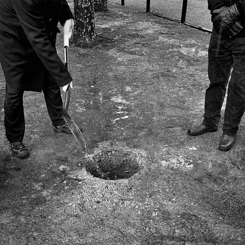 A person in a black coat shovels sand into a hole, while another person observes nearby in a graveyeard setting.