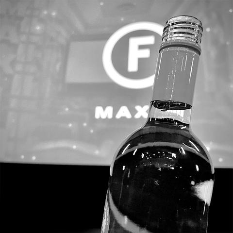 A close-up of a wine bottle in black and white, with a blurred partly obscured logo in the background showing the letter F in a circle and the text "MAX".