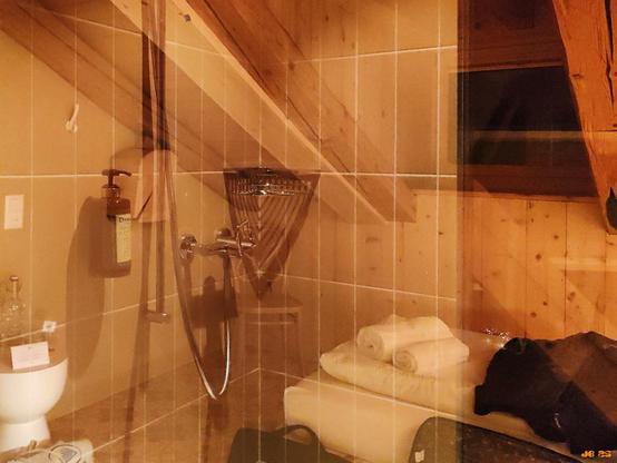 Farbfoto in gedämpften Farben im Orangebereich, das einen Blick in das kleine Bad eines Hotelzimmers zeigt, teilweise von Spiegelungen überlagert von der gegenüberliegenden Zimmerseite überlagert.