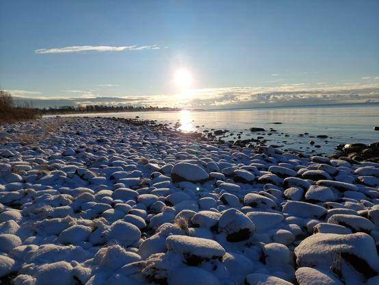 Photo of a snowy shore of round rocks, calm sea, blue skies and some clouds in the distance.