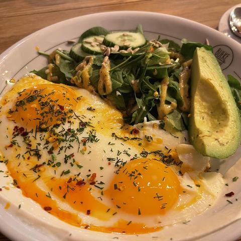 Na talerzu leżą dwa jajka sadzone z płynnymi żółtkami, posypane koperkiem i płatkami chili. Obok znajduje się garść rukoli z ogórkiem i pestkami słonecznika polana musztardowym sosem oraz połówka awokado. Całość podana na jasnym talerzu na drewnianym stole.