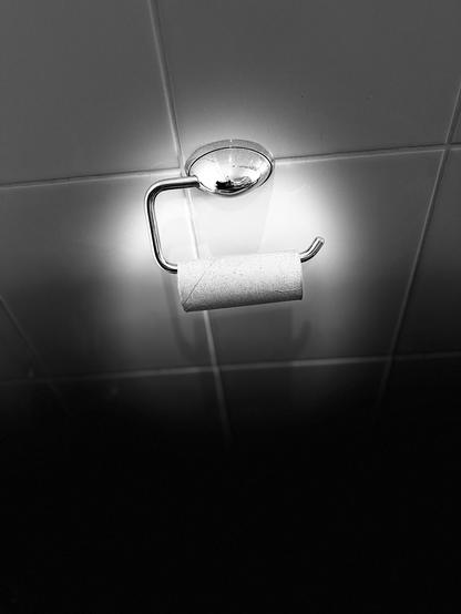 Nahaufnahme eines leeren Toilettenpapierhalters, der an einer Wand befestigt ist, beleuchtet von einer Deckenlampe, vor einem dunklen Hintergrund. Das Bild ist in Schwarz-Weiß.