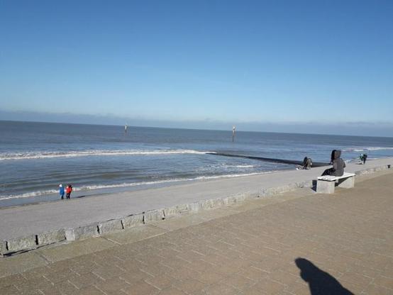 Im Vordergrund der mit Pflastersteinen befestigte Deich, im Hintergund die Weite des blauen Meeres mit leichten Wellen. Der Himmel strahlend hell bis dunkelblau. Eine Person sitze in dicker Jacke auf einer Bank aus hellem Stein, den Rpcken zum Fotografen, mit dessen Schatten am unteren Bildrand. Mehrere Personen in bunten Jacken spazieren am Spülsaum