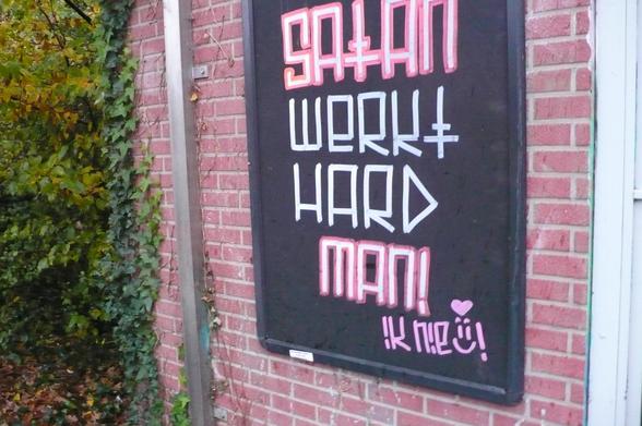 stenen muurtje met struiken begroeiing, een metalen lat en rechts het kozijn van een deur. In het reclame frame een zwarte poster met roze met witte letters