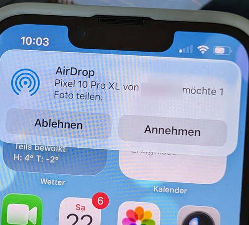 Ein Bild eines Smartphone-Bildschirms zeigt ein Pop-up-Fenster über AirDrop. Das Pop-up-Fenster hat einen hellblauen Hintergrund mit abgerundeten Ecken. Oben steht "AirDrop" und darunter "Pixel 10 Pro XL möchte ein Foto teilen". Unterhalb davon befinden sich zwei Schaltflächen: "Ablehnen" auf der linken Seite und "Annehmen" auf der rechten Seite. Im unteren Bereich des Bildschirms befinden sich App-Symbole, darunter "Wetter" und "Kalender", mit einer Benachrichtigungsblase mit der Zahl "6" über dem Kalender-Symbol. Oben auf dem Bildschirm sind die Uhrzeit "10:03" und Symbole für Mobilfunk- und WLAN-Verbindung angezeigt.