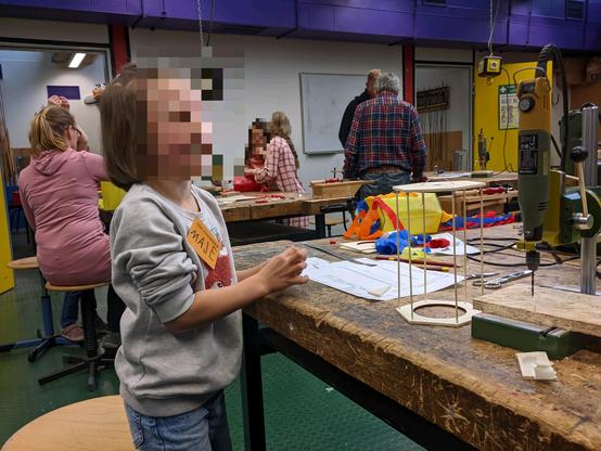 Werkbänke mit kleinem Bohrschleifer im Bohrständer. Auf dem Tisch im Vordergrund das Grundgerüst einer Lampe aus Holz. Mehrere Kinder und Erwachsene beim Arbeiten.