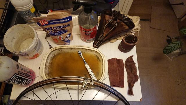 Jante, étriers de freins et selle d'un vélo ancien après traitement chimique et mécanique de la rouille. Avec de droite à gauche : 
- paille de fer fine & bande de papier à poncer type émeri haute résistance pour métaux (KK711X)
- selle de vélo après un rapide traitement de la rouille
- vinaigre blanc 12°
- sachet d'acide citrique (en cours d'utilisation dans la bassine)
- seau d'eau
- pétrole 
- nettoyant à vélo