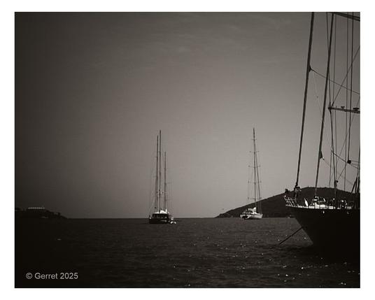 Black and white photo of sailboats anchored in calm waters. Two boats are centered with tall masts, while another is silhouetted on the right. A peaceful, nostalgic tone.