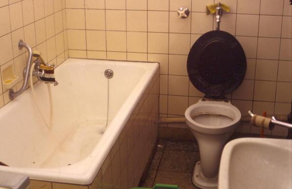 Foto eines alten Badezimmers. Eierschalenfarbene kleine Wandfliesen, Badewanne mit Schmutzrändern, Toilette mit schwarzem geöffnetem Deckel, Waschbecken. Über dem Handtuchhalter hängt eine leere Klopapierrolle.