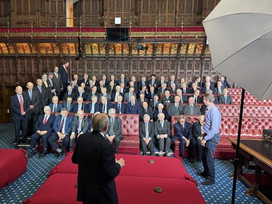 The image depicts a large group of people seated in a richly decorated hall with dark wood paneling and blue patterned carpeting. The hall appears to be an assembly room, possibly a parliament or government building. Most of the individuals are seated in rows of red upholstered chairs, facing towards the left of the frame, with several people standing near the right side. The people are wearing suits, mostly dark in color, with some wearing ties. The hall features ornate detailing throughout, including intricate patterns on the walls and ceiling, and a few circular green objects on the floor near the front of the room.
