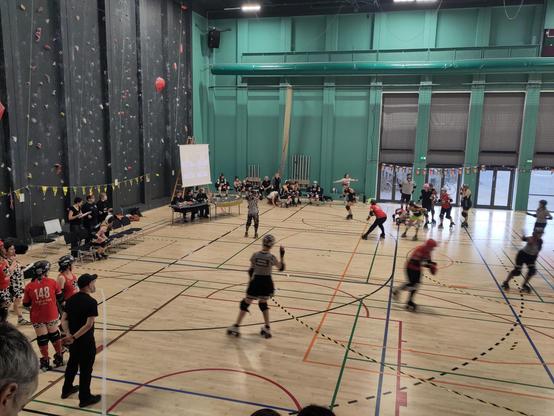 Gym hall. A lot of people o scates. Some on chairs. Some in a pack on the floor. Half in red cloth, half in yellow/black cloth.