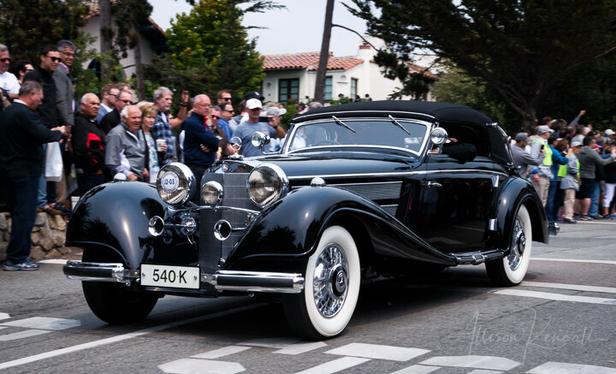 A classic old car drives down the main avenue of a small coastal California town. Crowds of spectators watch from the sidewalk. The car is a large vintage Mercedes with dramatic curves and chrome details.