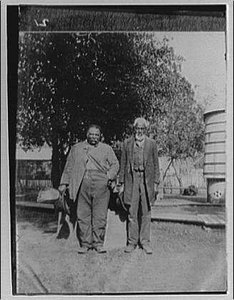 The image is a black and white photograph featuring two men standing side by side on what appears to be a paved area, possibly in an urban environment. The man on the left wears overalls with rolled-up sleeves and holds a gardening tool or implement that resembles a hoe near his right hand. His posture suggests he may have been working recently.

The second man stands taller than the first; dressed in trousers, shoes, a shirt, waistcoat, jacket, and possibly glasses (the style of which is not clearly visible due to image quality). He has a mustache, beard, and appears older with his head slightly tilted forward. His right hand seems relaxed at his side.

Behind them stands an old-fashioned building or structure with what could be windows in the upper part, suggesting this might have been taken near buildings used for various purposes such as shops, housing, etc., typical of early 20th-century urban settings.

A tree is visible behind these men providing partial shade to their right side. The background also includes another large object which looks like a cylindrical structure that could be an old water tower or similar industrial building.

The overall composition suggests this photograph captures a moment in everyday life from around the time it was taken, offering glimpses into social and architectural contexts of its era.