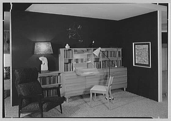 The image depicts a monochrome photograph of an interior room, likely from the mid-20th century. The space features dark walls and is furnished with modernist-style furniture including a large desk or credenza against one wall adorned with numerous books on shelves. Above this piece of furniture hangs abstract art pieces that resemble wire sculptures in white.
Adjacent to the book-filled shelf is an armchair, upholstered in patterned fabric, possibly velvet. A solitary lamp sits atop it, its light casting onto nearby surfaces and creating a warm ambiance. To one side stands another chair, with a visible backrest suggesting minimalist design principles at play within this setting.

On the wall opposite the furniture ensemble hangs what appears to be an area rug or decorative textile patterned in squares of various shades. The room is also decorated with additional artworks including framed maps and abstract shapes which contribute to its contemporary aesthetic.
The flooring seems carpeted, adding a layer of texture beneathfoot while maintaining coherence within this space's design scheme.

Overall, the image captures an elegantly arranged interior designed with modernist sensibilities in mind, showcasing both functional elements like storage spaces and decorative aspects such as art pieces that reflect mid-century tastes.