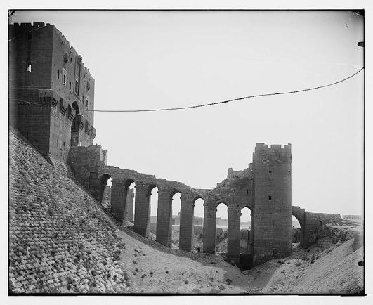 The image displays a historical castle ruin with remnants of towering walls and archways. The structure appears aged, made primarily of stone blocks, exhibiting signs of decay such as missing stones or collapsed sections. It's set against a barren landscape under an overcast sky, suggesting arid conditions possibly in the middle eastern region based on architectural style. There is minimal vegetation, with sparse patches of grass and shrubs around the base of the ruins. The photograph captures this scene from a perspective that looks up at the castle remains, emphasizing their height and grandeur despite deterioration.