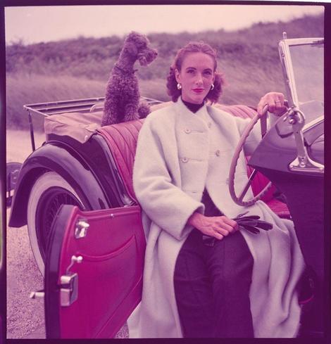 A woman sits in the driver's seat of an old-fashioned convertible automobile, with a red leather suitcase at her feet and holding what appears to be a pair of gloves. She is wearing a light-colored coat over dark trousers and has styled hair adorned with accessories. A poodle dog sits on a pink upholstered luggage rack behind her, gazing out into the distance. The backdrop features an open field or dune area under a cloudy sky. This image appears to be part of Toni Frissell's collection of portraits from the 1940s-1950s, particularly capturing American life and coastal scenes along New England beaches.